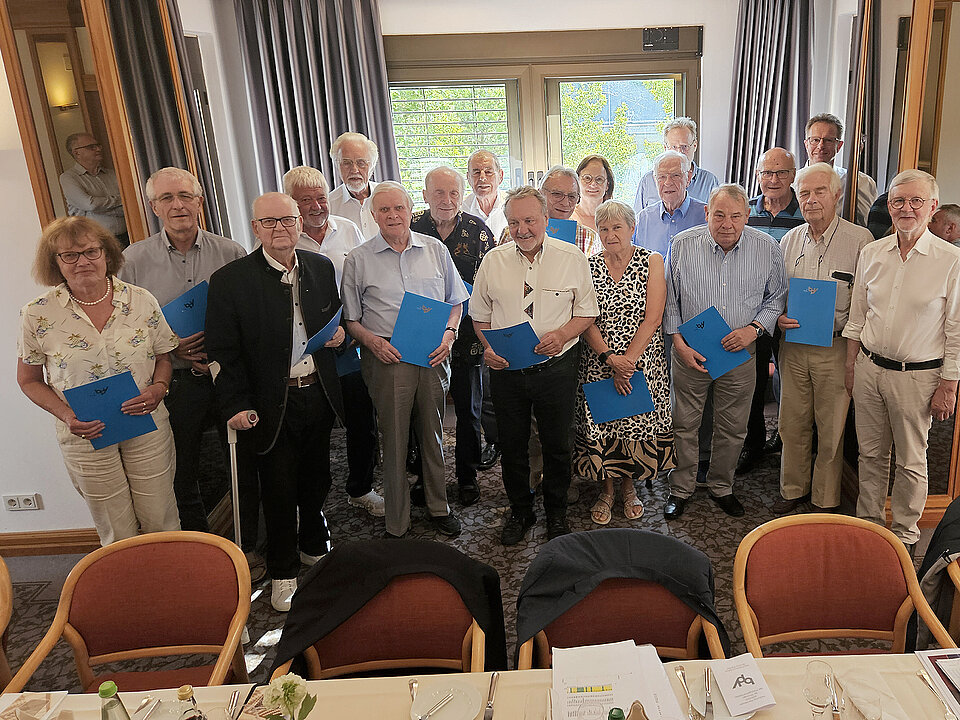 Eine Gruppe von Seniorinnen und Senioren steht in einem festlich eingerichteten Raum und hält blaue Ordner, während vor ihnen ein gedeckter Tisch mit Getränken und Besteck steht. Beschreibung von KI generiert
