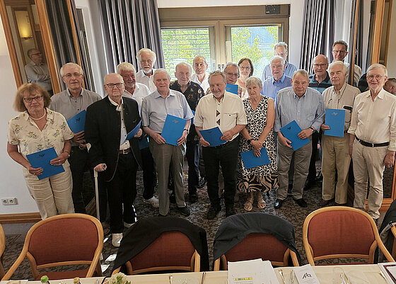 Eine Gruppe von Seniorinnen und Senioren steht in einem festlich eingerichteten Raum und hält blaue Ordner, während vor ihnen ein gedeckter Tisch mit Getränken und Besteck steht. Beschreibung von KI generiert