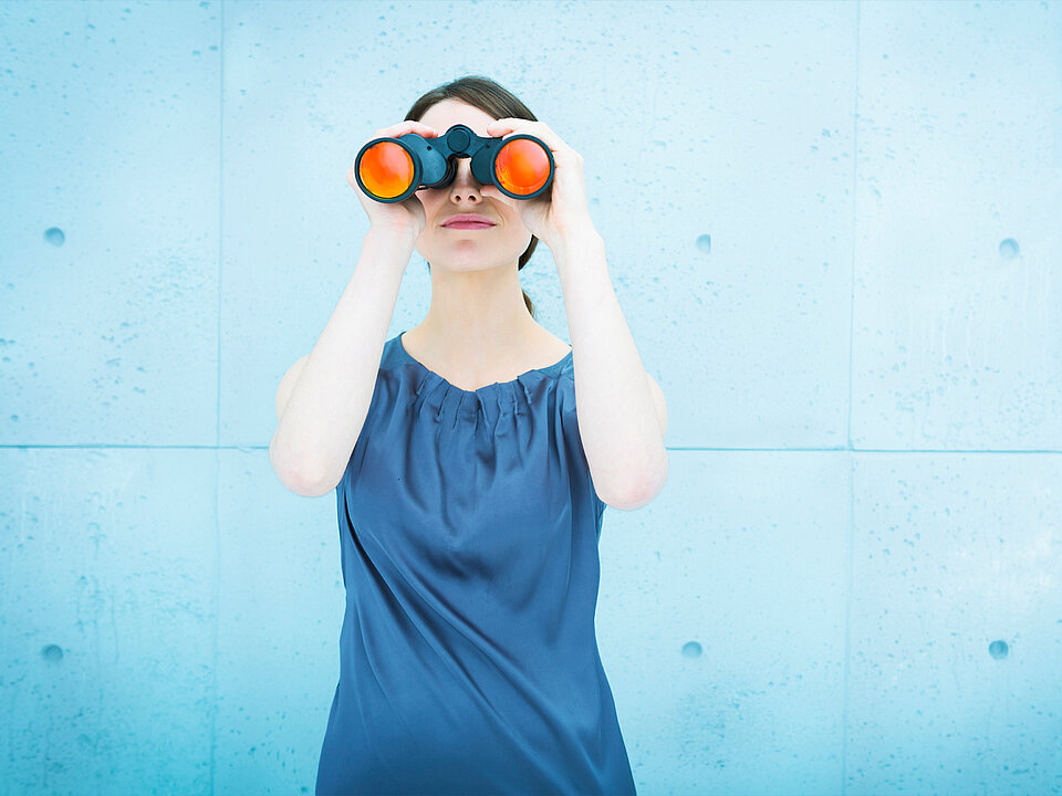 Eine Person mit kurzen dunklen Haaren hält ein Fernglas mit orangefarbenen Gläsern vor die Augen vor einem hellblauen Hintergrund. Beschreibung von KI generiert