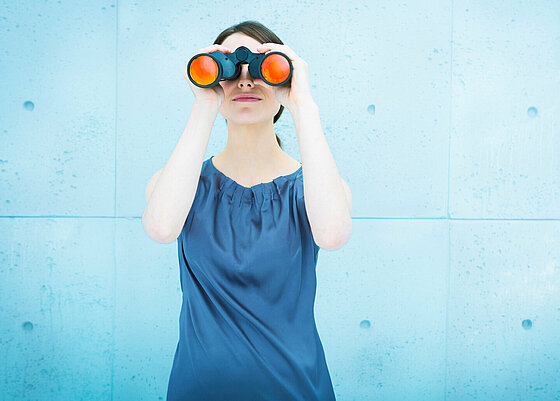 Eine Person mit kurzen dunklen Haaren hält ein Fernglas mit orangefarbenen Gläsern vor die Augen vor einem hellblauen Hintergrund. Beschreibung von KI generiert