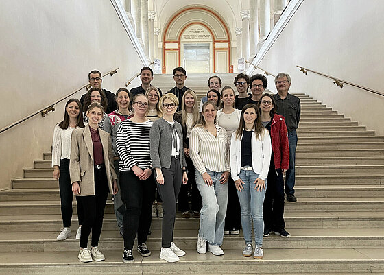 Eine Gruppe von Erwachsenen steht auf einer breiten Treppe in einem historischen Gebäude mit hohen Wänden und einem Bogen im Hintergrund. Beschreibung von KI generiert
