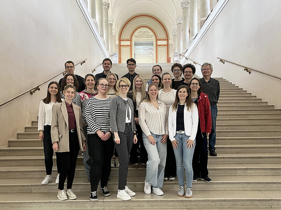 Eine Gruppe von Erwachsenen steht auf einer breiten Treppe in einem historischen Gebäude mit hohen Wänden und einem Bogen im Hintergrund. Beschreibung von KI generiert