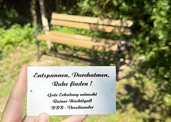 Eine Hand hält eine Karte mit einem Text vor einer Parkbank und grünen Bäumen im Hintergrund. Beschreibung von KI generiert