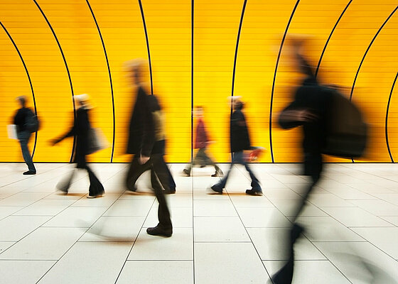Mehrere Menschen in Bewegung sind vor einer gelben Wand mit schwarzen Linien in einem modernen Innenraum zu sehen. Beschreibung von KI generiert