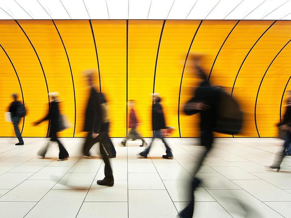 Mehrere Menschen in Bewegung sind vor einer gelben Wand mit schwarzen Linien in einem modernen Innenraum zu sehen. Beschreibung von KI generiert