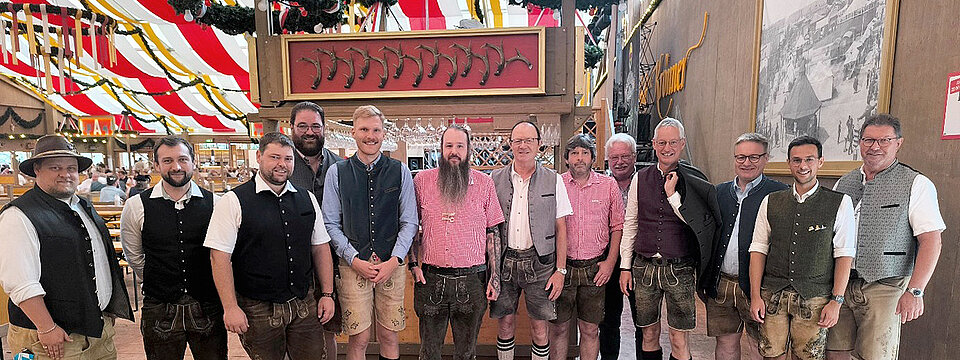 Eine Gruppe von Erwachsenen trägt traditionelle bayerische Trachten und steht in einem festlich dekorierten Zelt mit rot-weißen Stoffbahnen und Holzverzierungen. Beschreibung von KI generiert