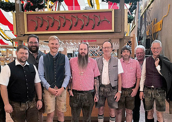 Eine Gruppe von Erwachsenen trägt traditionelle bayerische Trachten und steht in einem festlich dekorierten Zelt mit rot-weißen Stoffbahnen und Holzverzierungen. Beschreibung von KI generiert