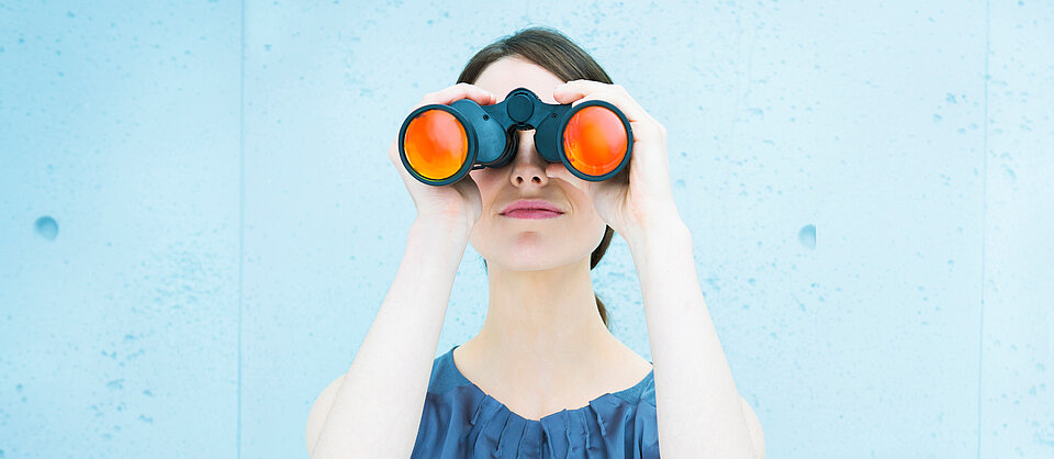 Eine Person mit kurzen dunklen Haaren hält ein Fernglas mit orangefarbenen Gläsern vor die Augen vor einem hellblauen Hintergrund. Beschreibung von KI generiert